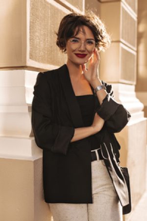 Joyful lady in jacket and white pants smiling outside. Lovely woman with bright lips in eyeglasses looking into camera outdoors.